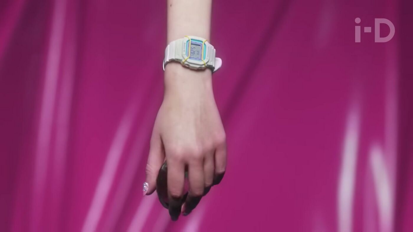 A white Baby-G with yellow protective wires appears on a wrist against a pink background.