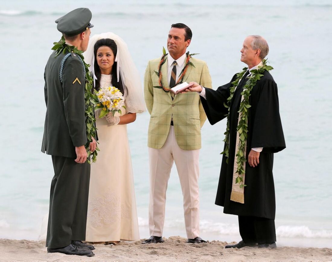 Don stands beside a bride, groom and officiant at a wedding on the beach.