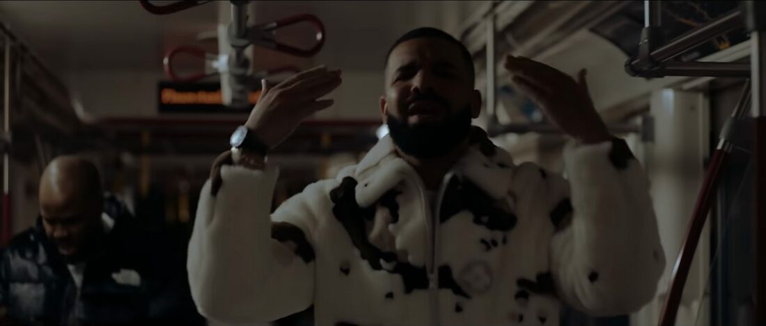 Drake raises both hands in a subway car, with light reflecting from his watch.