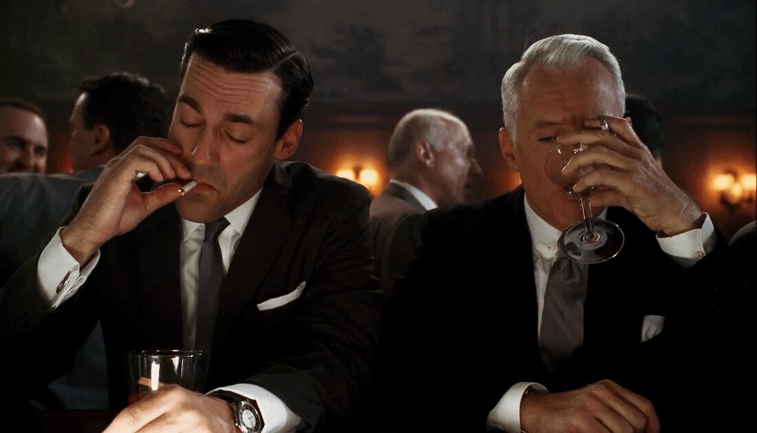 Don sits beside Roger at a table, with a steel bracelet watch visible at his left cuff.