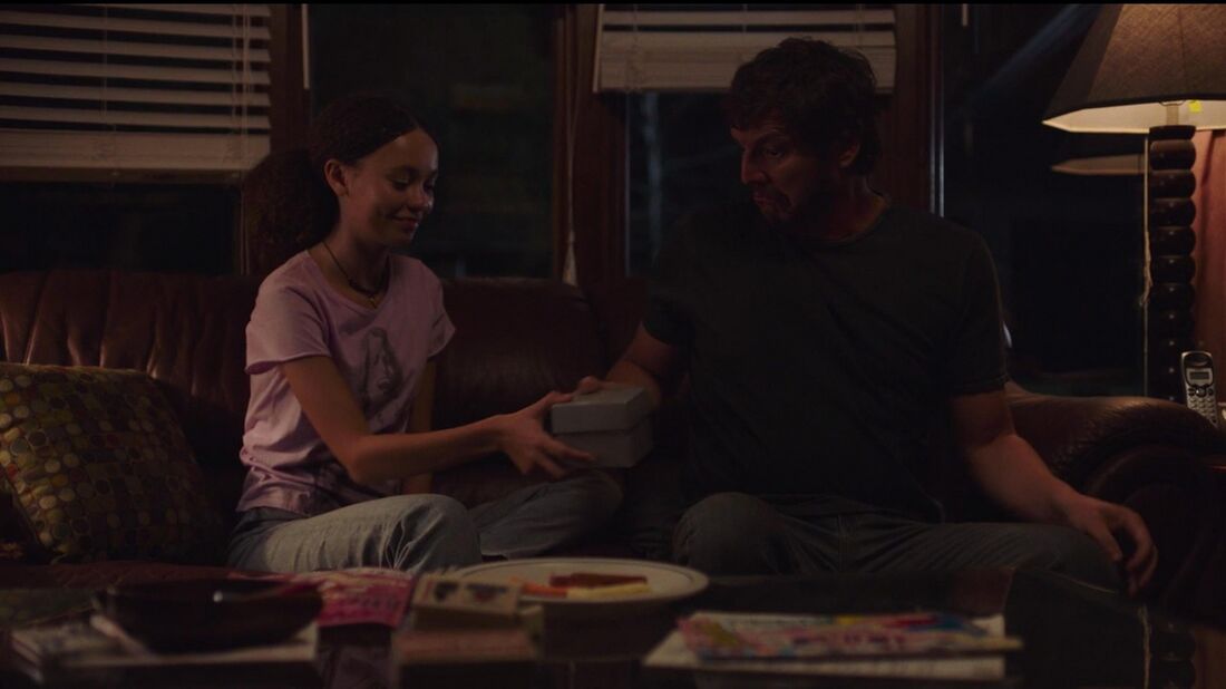 Sarah hands a gray gift box to Joel as they sit together on a couch.