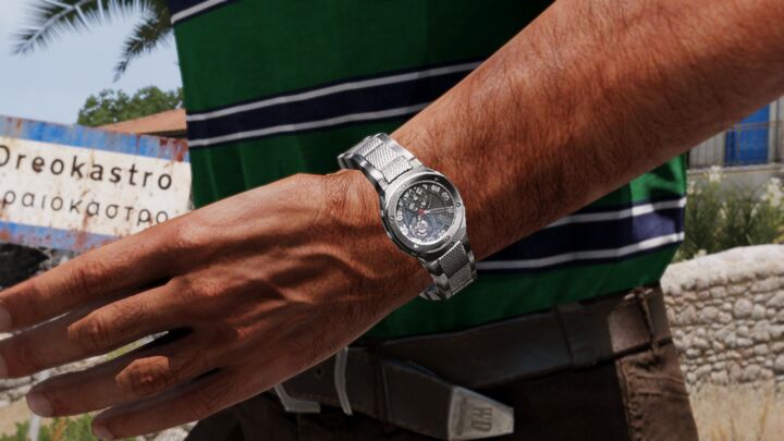 A steel-colored cushion-case watch with an asymmetric dial on a character’s wrist.
