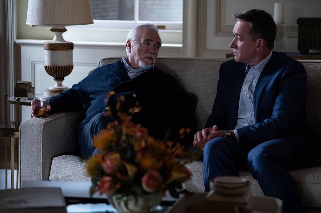 Tom and Logan sit on a sofa, with Tom’s steel watch bracelet visible above his clasped hands.