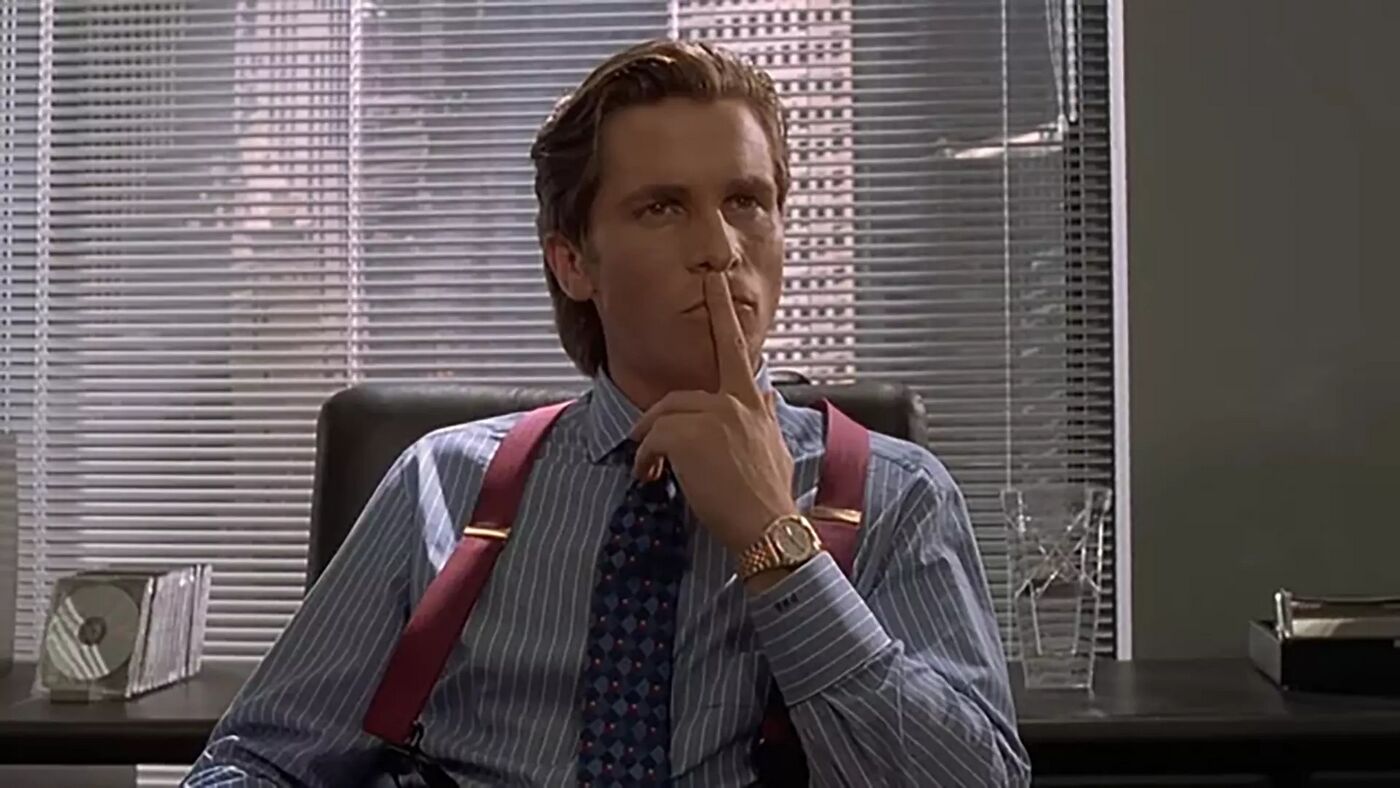 Christian Bale as Patrick Bateman raising a finger to his lips while wearing a two-tone watch in American Psycho