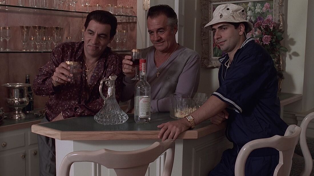 Christopher wears a navy tracksuit and bucket hat beside Silvio and Paulie, with a rectangular gold watch at his wrist.