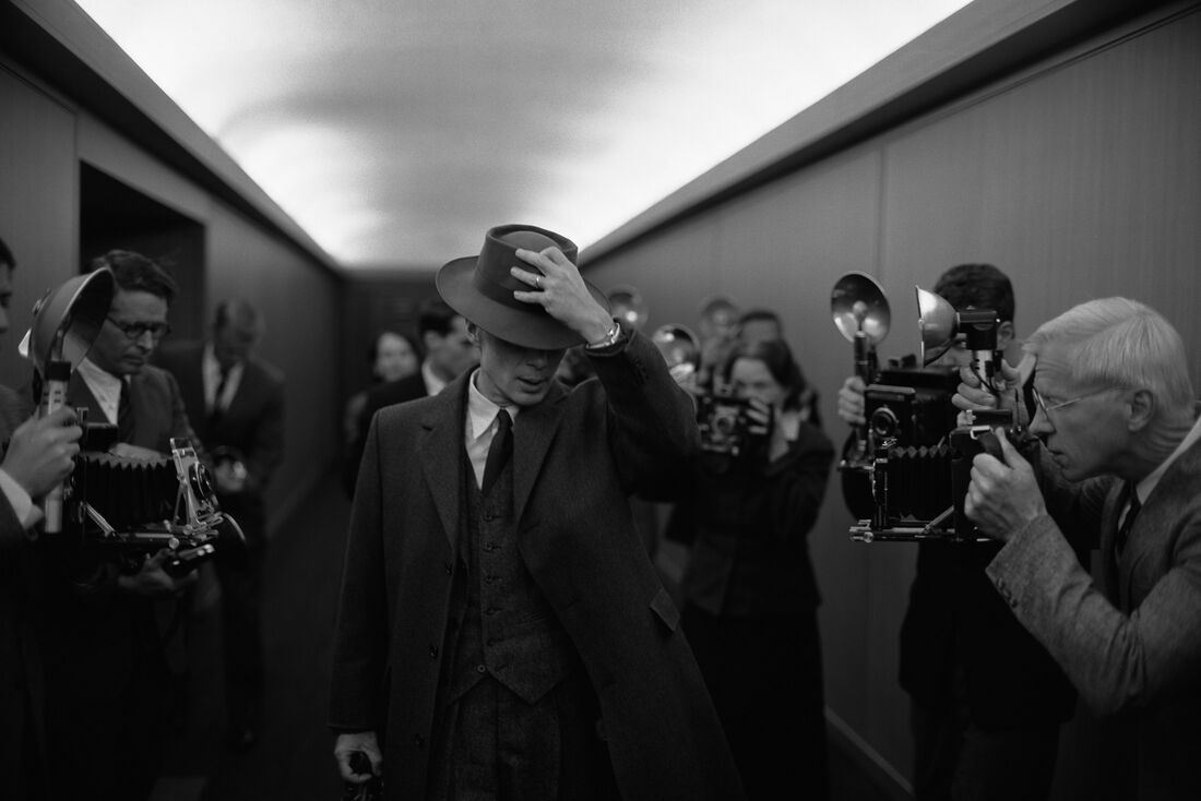 Cillian Murphy as J. Robert Oppenheimer tipping his hat in a press corridor, a vintage Hamilton on his raised left wrist