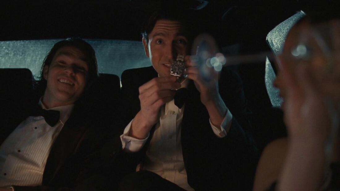 Greg holds a steel bracelet watch in front of him while another man watches.