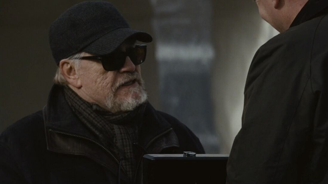 Logan, wearing a black cap and sunglasses, looks up toward Tom while holding the presentation box.